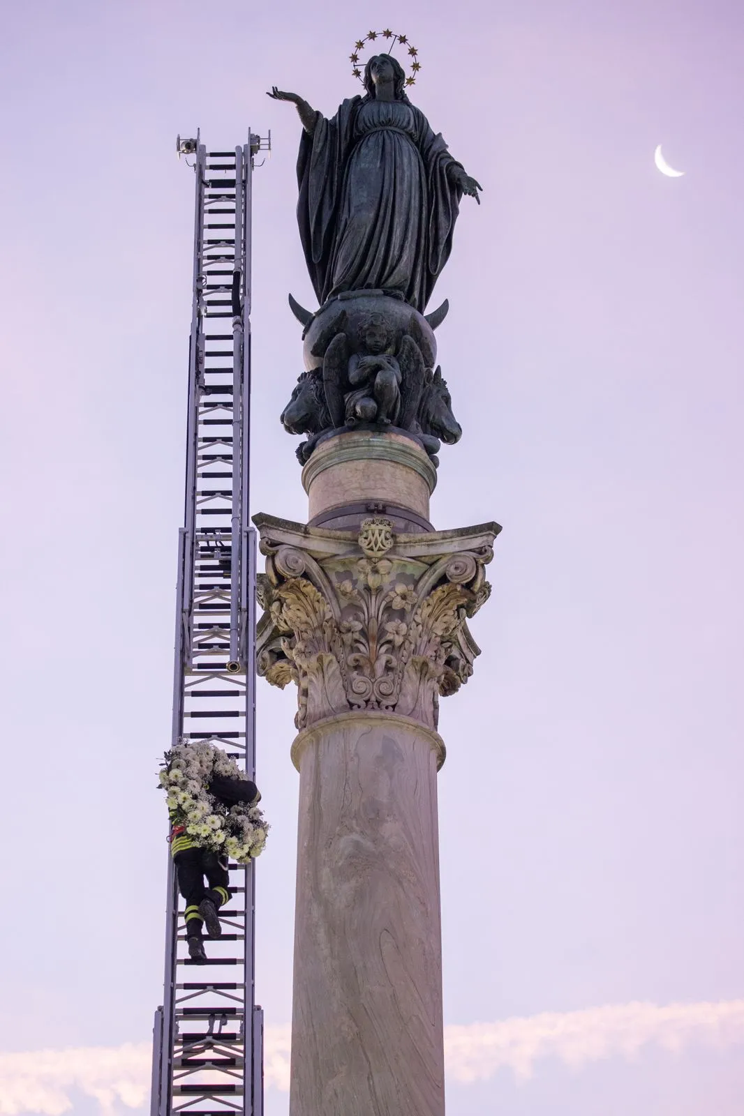 A firefighter scales a long ladder to the top of a nearly 40-foot-high column to pay tribute at dawn to the Blessed Virgin with a wreath of flowers on Dec. 8, 2023. Credit: Daniel Ibaniez/CNA